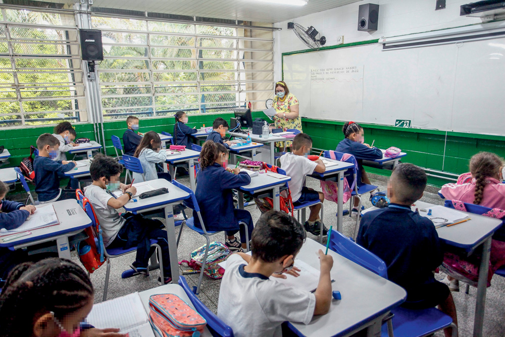 Crianças sentadas em sala de aula, escrevendo em suas mesas. Ao fundo, professora aparece sentada em sua mesa