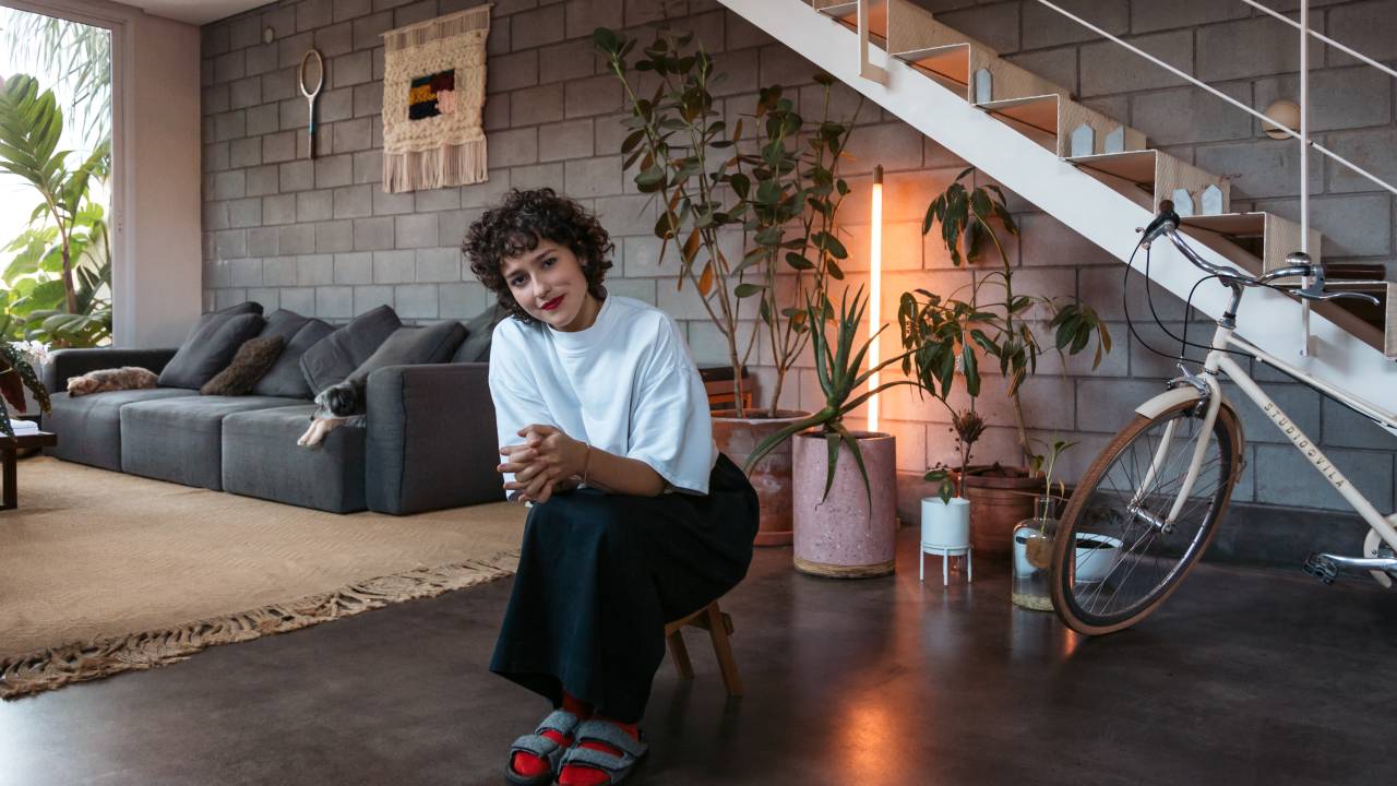 Bebel Luz posa sentada em banquinho de madeira em sala com escadas, plantas e sofá cinza ao fundo.