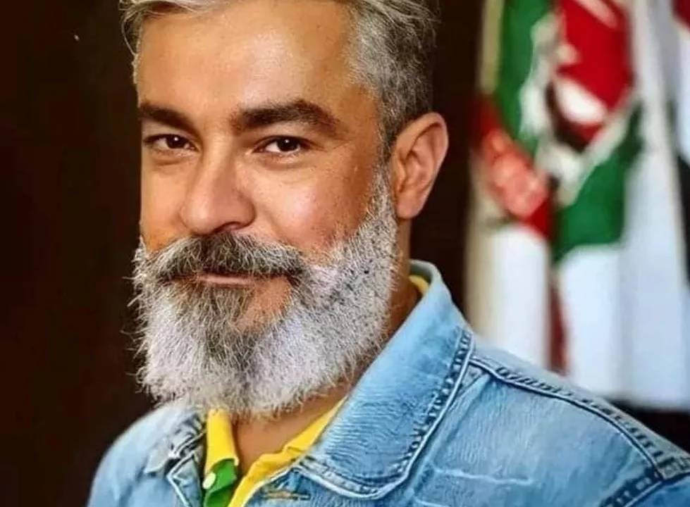 Foto exibe homem com barba e cabelo grisalhos vestindo jaqueta jeans clara e camisa amarela.