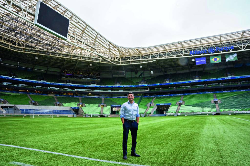 Imagem mostra homem de camisa azul e calça preta no meio de campo de futebol em estádio