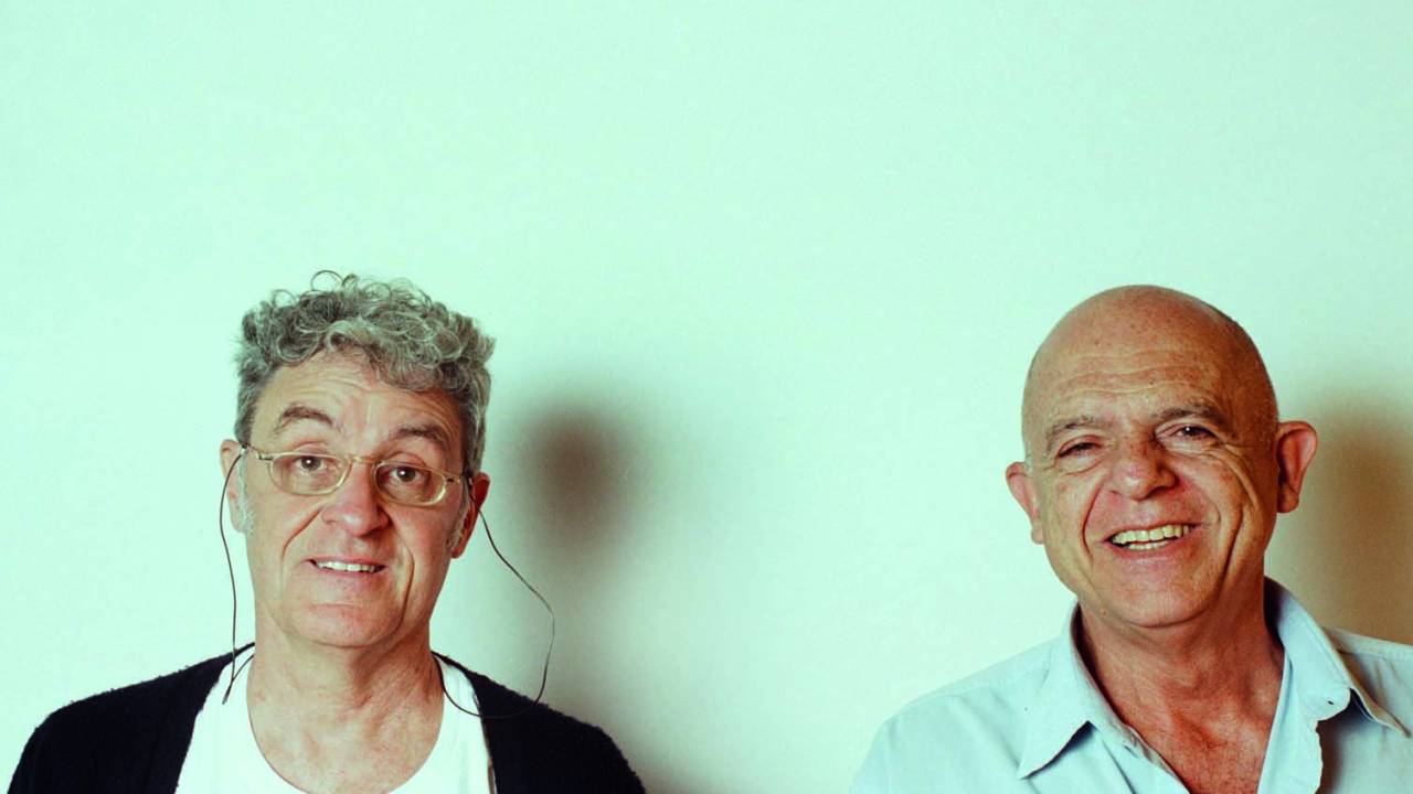 Imagem mostra dois homens sobre fundo branco, sorrindo, olhando para a câmera