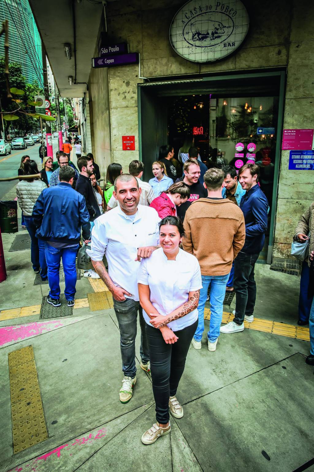 Imagem mostra casal de chefs vestindo roupas brancas em esquina com aglomeração na entrada de estabelecimento