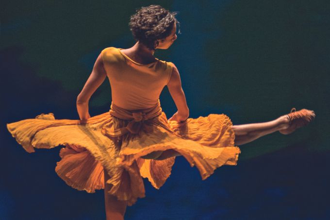 Dançarina branca, magra, de cabelos escuros, curtos e cacheados, dá um rodopio no palco. Ela é vista de costas e usa um vestido amarelo esvoaçante