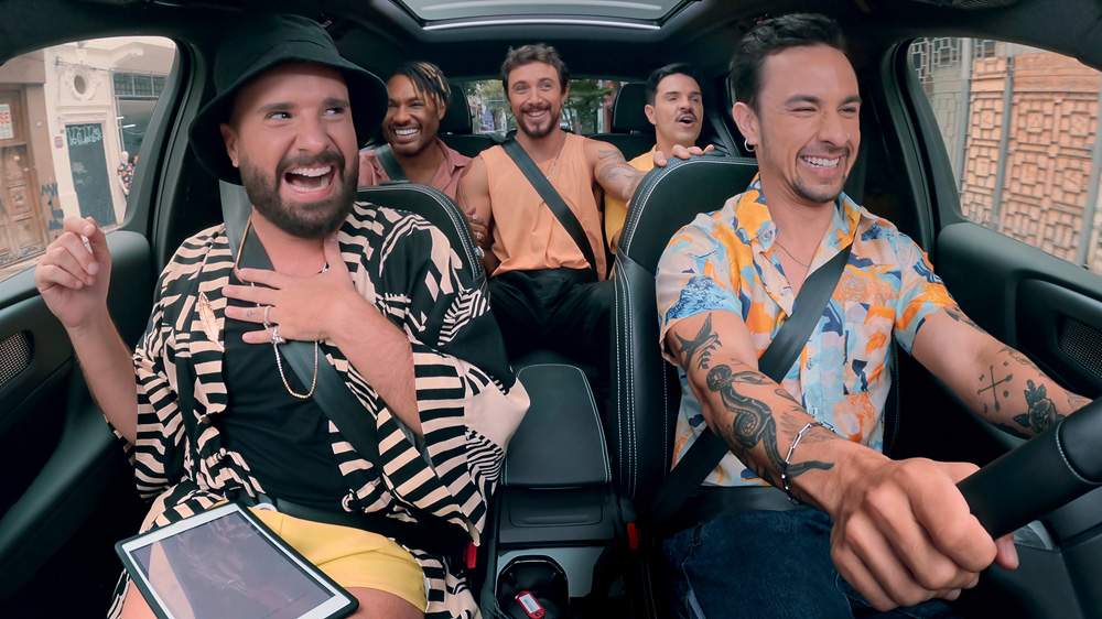 Imagem mostra cinco homens sorrindo dentro de carro, vestindo roupas de tons amarelos