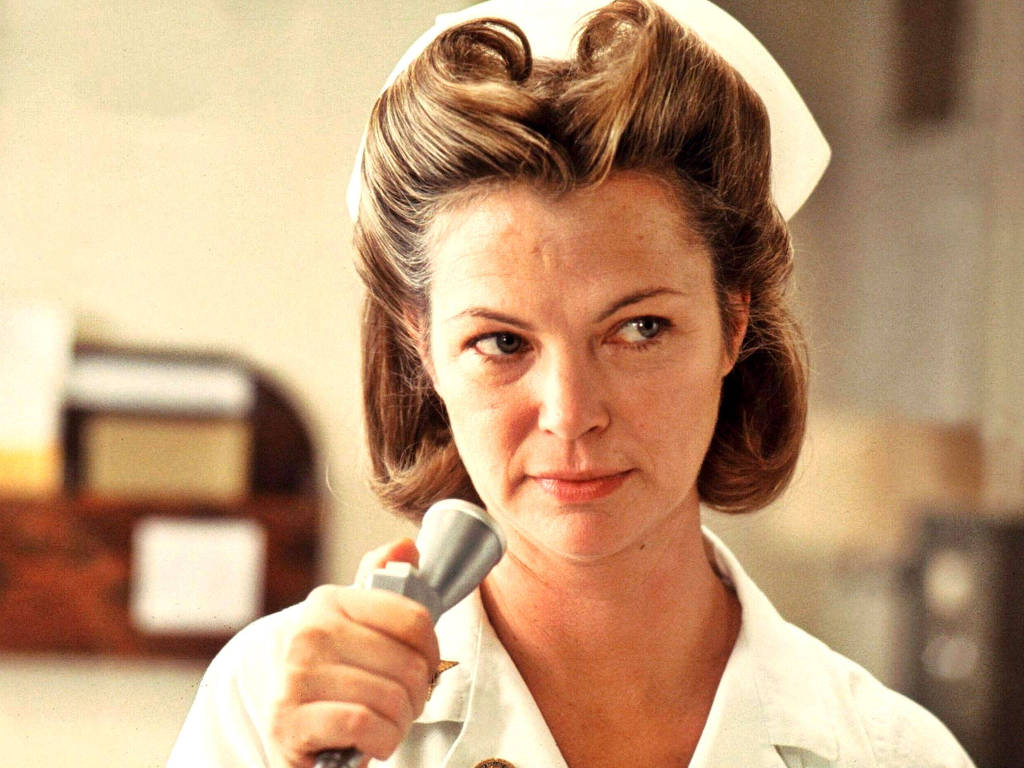 Louise Fletcher vestida de enfermeira segurando um microfone prateado.