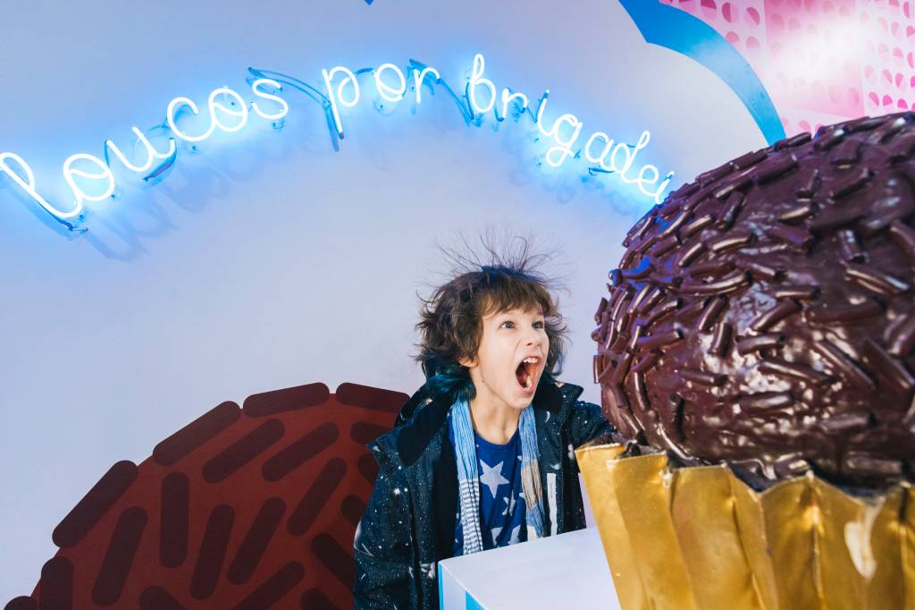 Brigadeiro – Lana Pinho Criança grita em frente a brigadeiro gigante. Ao fundo, neon azul diz