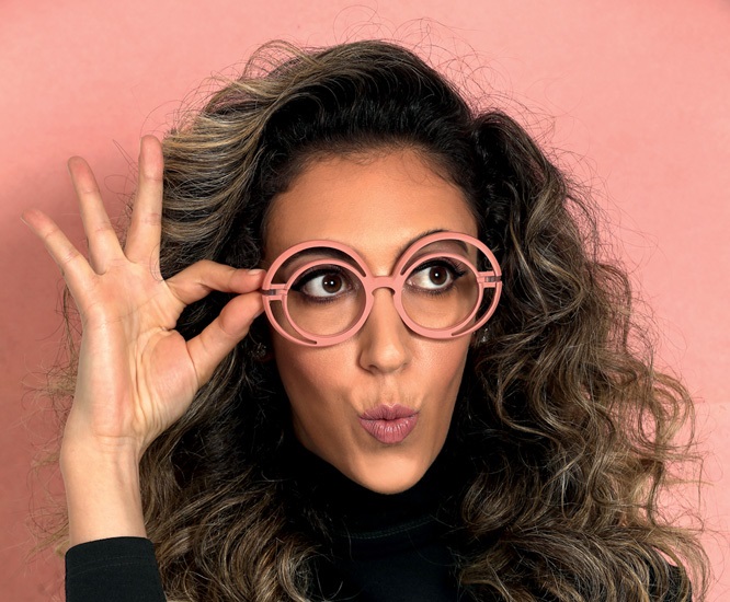Chantal olha para a direita com uma das mãos segurando os óculos ovais cor-de-rosa no rosto. Ela veste suéter preto e tem os cabelos encaracolados e quase pretos.
