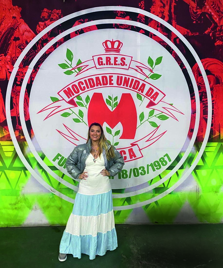 Imagem mostra mulher com vestido branco e azul, sorrindo em frente a cartaz de escola de samba