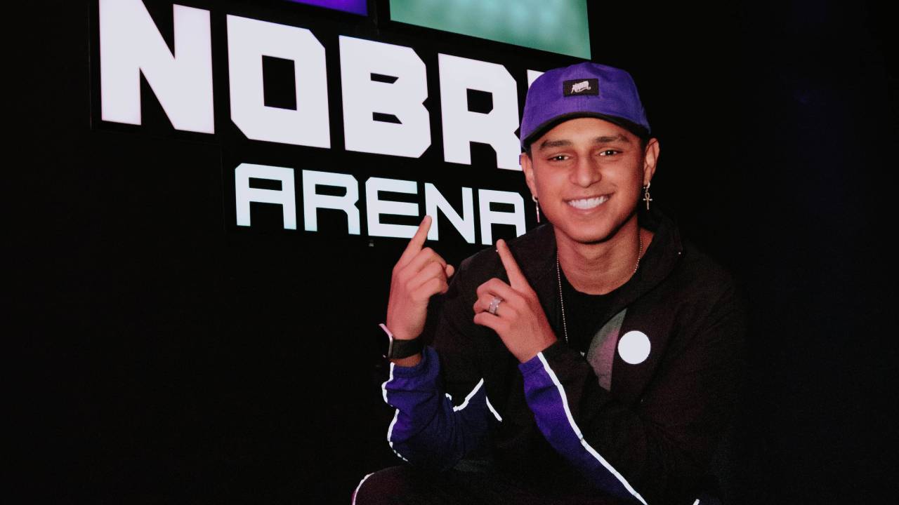 Gamer Nobru posa agachado, sorrindo e apontando para placa que está às suas costas, escrito Nobru Arena