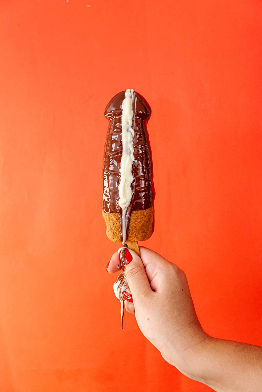 Waffle em formato de pênis fotografado de frente com mão segurando-o. Aparece banhado na cor marrom de chocolate e uma gota branca escorrendo inteira.