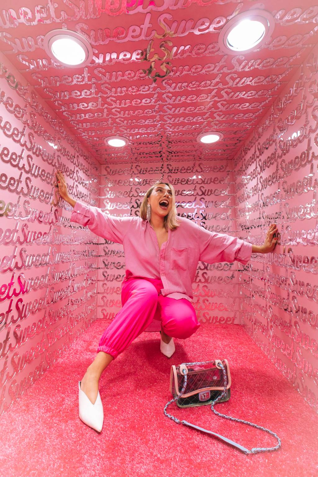 O museu mais doce do mundo – Lana Pinho-20 Moça vestida toda de rosa aparece agachada em cenário rosa com bolsa preta no chão. Ela está de braços abertos e tem cabelos loiros.