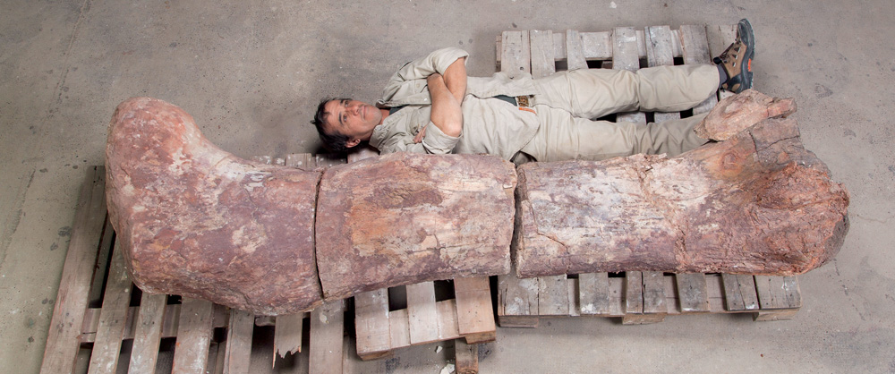 TERCEIRA Crédito_ J.M. Farfaglia_Divulgação.jpg Técnico de macacão bege deitado ao lado de um fóssil gigante de fêmur de dinossauro