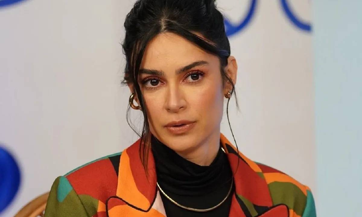 Atriz Thaila Ayala posa diante de fundo neutro, com semblante neutro, usando camiseta marrom e laranja