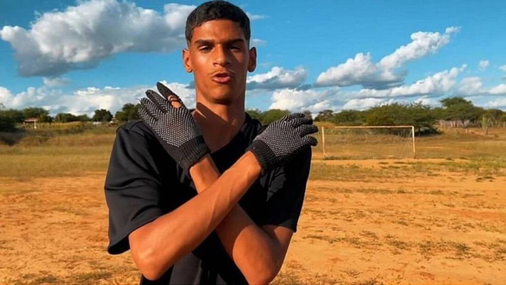 Luva de Pedreiro posa cruzando os braços e fazendo um X, em campo de futebol de terra, com luva de goleiro