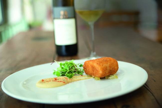 Prato branco raso contendo bacalhau, grão de bico e cidra cristalizado, com uma garrafa de vinho tinto ao fundo