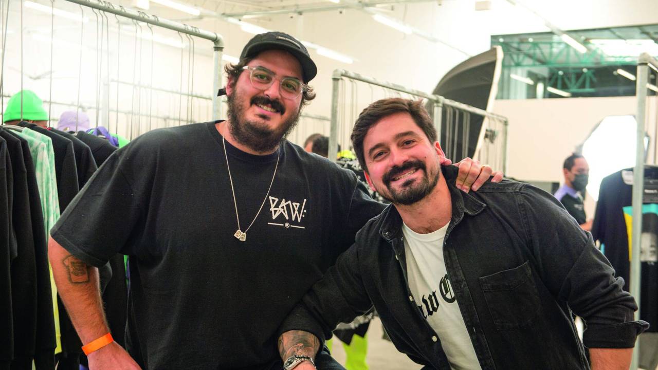 Dois homens posam lado a lado sorrindo em fábrica. Ambos vestem peças pretas de roupa.