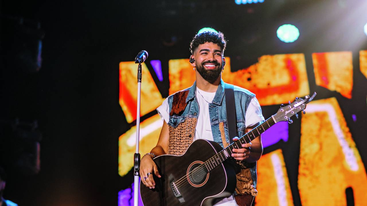 Foto de Dilsinho durante apresentação, segurando violão e sorrindo para a plateia