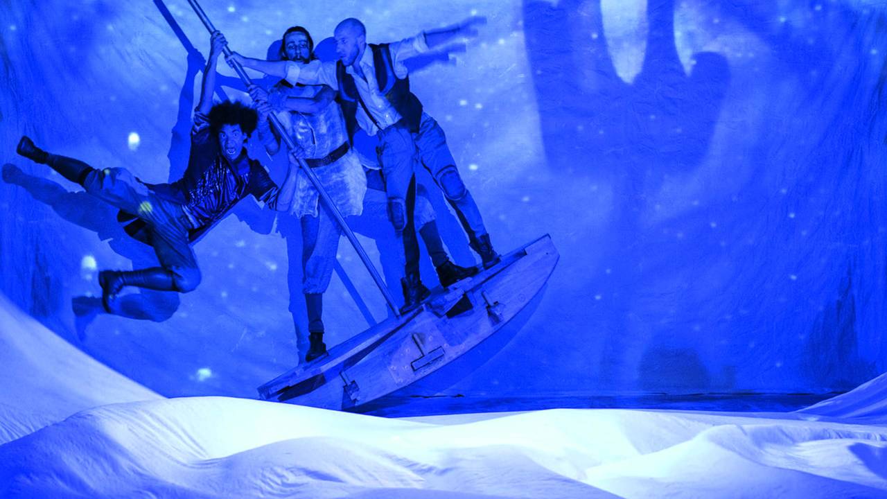 Três atores se balançam em barco de madeira em oceano cênico. Eles são iluminados por uma luz azul