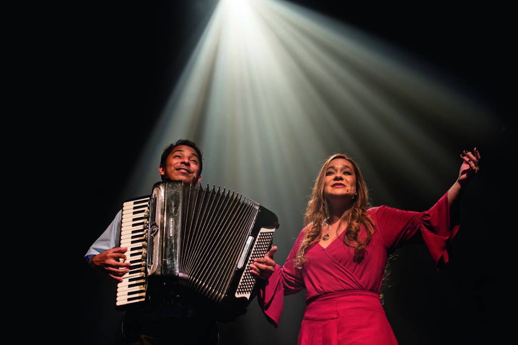 Cosme Vieira, homem negro, toca sanfona ao lado de Liv Moraes, mulher morena, que canta. Ela usa um vestido rosa escuro e está com os braços abertos