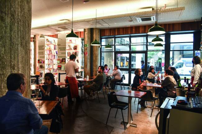Cuia Café ambiente Ambiente interno em que há mesas com pessoas sentadas, ao fundo estantes repletas de livros e à direita uma grande porta envidraçada que toma a parede toda