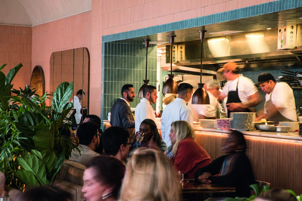 Ambiente La Serena com parede de azulejos rosados, abertura para a cozinha onde se encontram alguns garçons de branco e pessoas trabalhando no preparo dos pratos, uma planta no fundo, à esquerda e pessoas sentadas nas mesas do restaurante