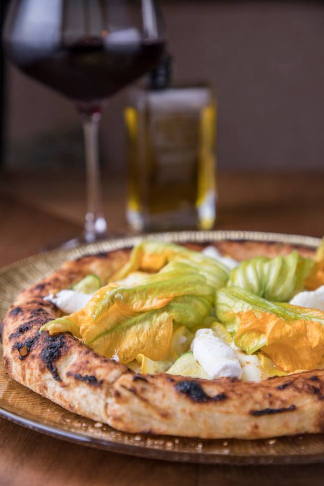 Massa de pizza individual assada coberta por flores amarelo esverdeadas dispostas sobre a massa, além de manchas de queijo muçarela fior di latte em volta delas