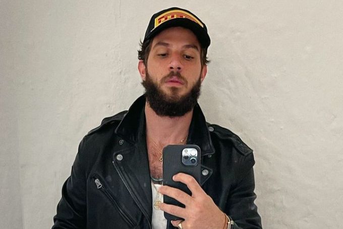 Selfie de Chay Suede no espelho, usando boné, jaqueta e camiseta branca, fundo branco