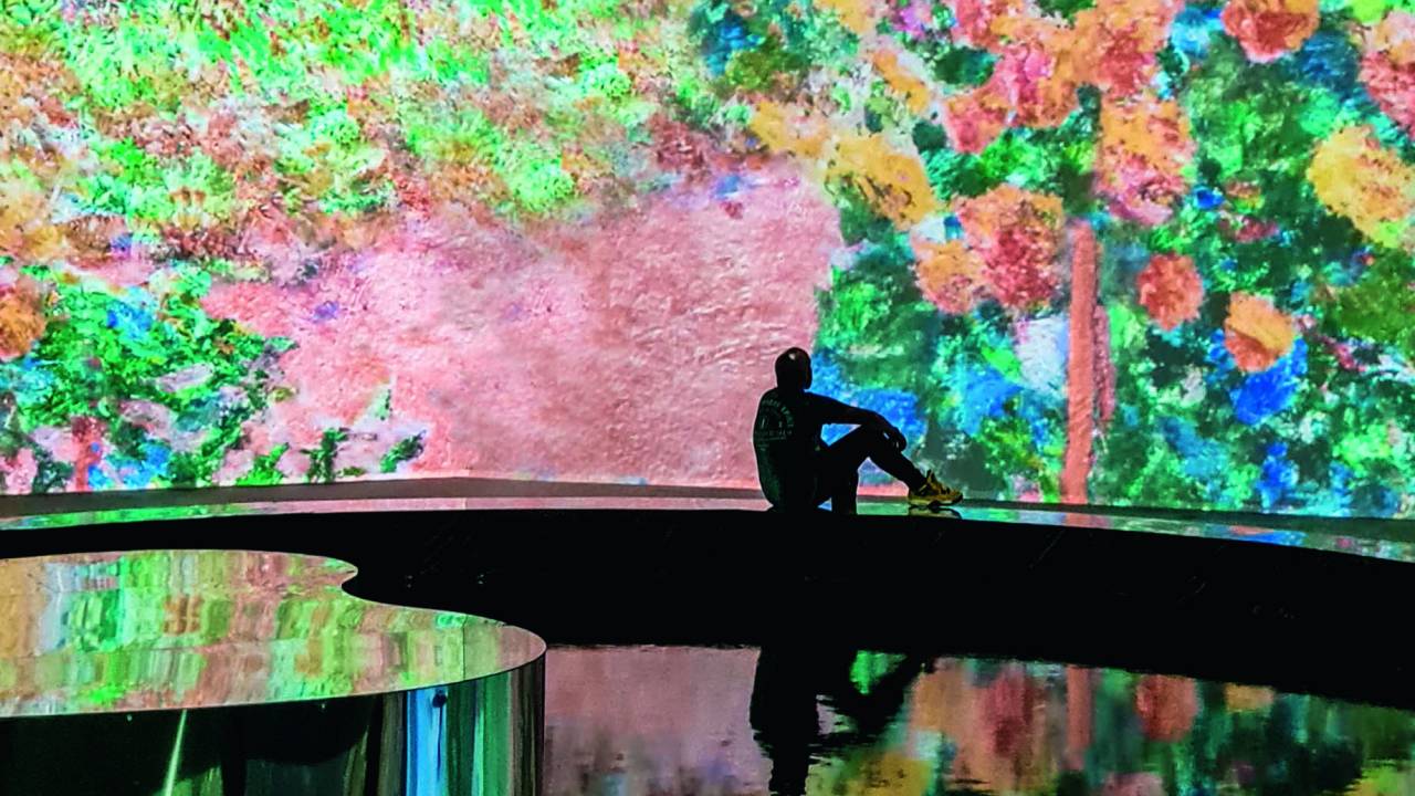 Visitante sentado em uma coluna circular e espelhada observa obra de Claude Monet projetada nas paredes e no chão