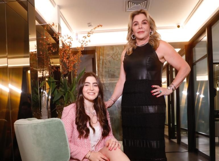 Valentina e Marisa Clermann posam lado a lado, a primeira sentada com blazer rosa e Marisa de pé com vestido preto.