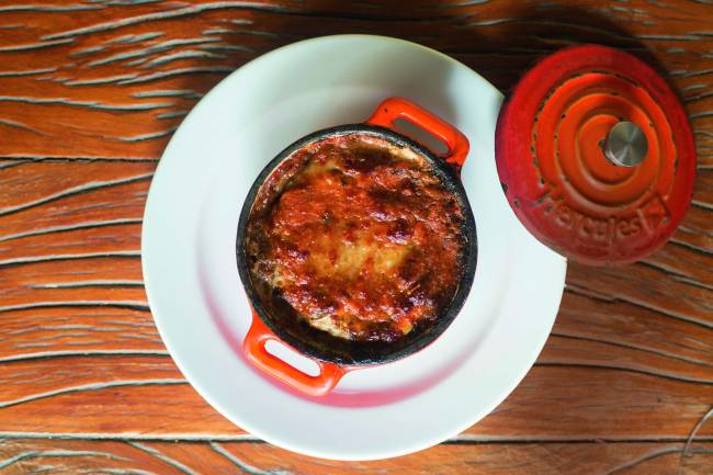 Panelinha de ferro vermelha contendo um parmigiana apoiada sobre um prato raso branco em uma mesa de madeira