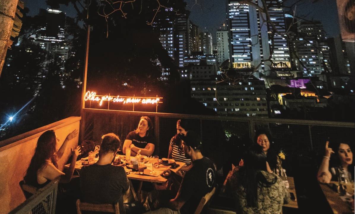Espaço externo com letreiro neon vermelho onde se lê olha pro céu meu amor, com pessoas reunidas conversando