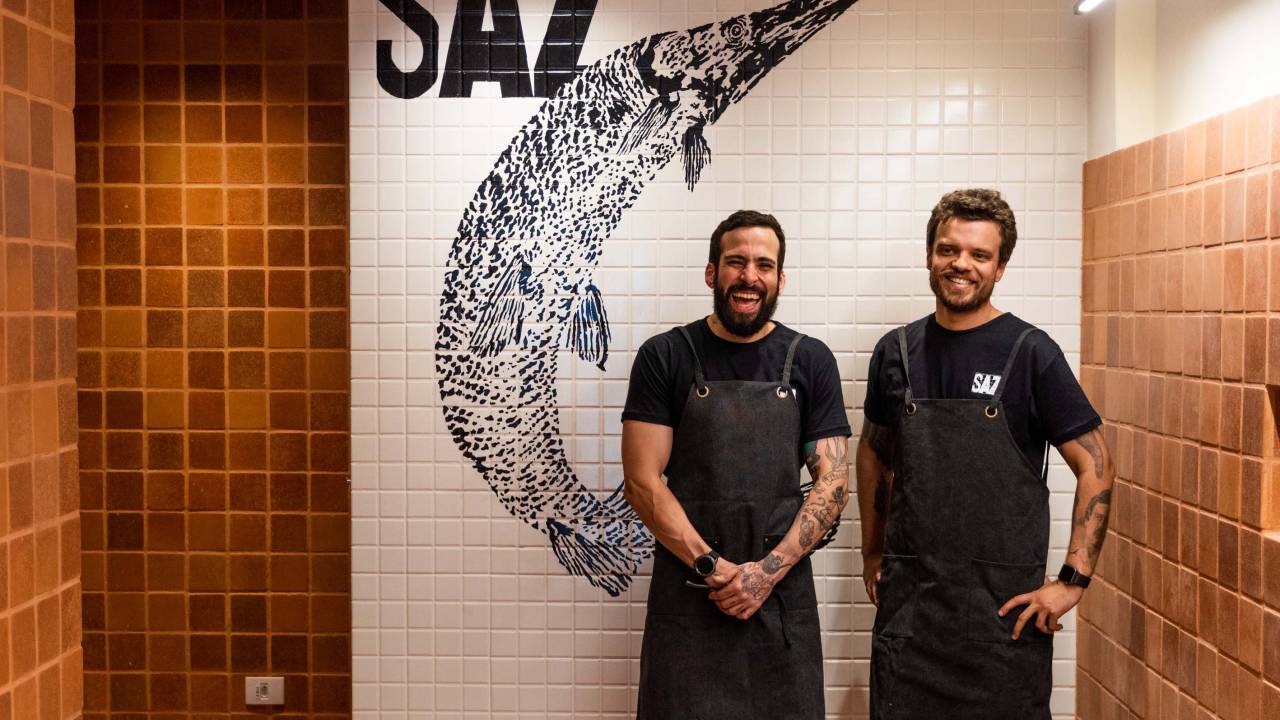 Tiellet de aventais e roupas pretas em frente a um painel de azulejos em que está desenhado um peixe e onde se lê "saz"