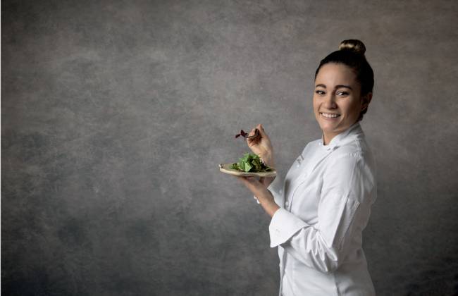 2022_08_28_Comer_e_Beber_Giovanna_Grossi_Wanezza_Soares-6984-Edit.psd Giovanna Grossi do Animus de pé à frente de uma parede cinza segurando um prato e um garfo e vestindo branco