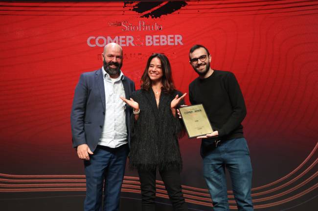 Mulher vestida de preto entre dois homens, um deles com uma placa