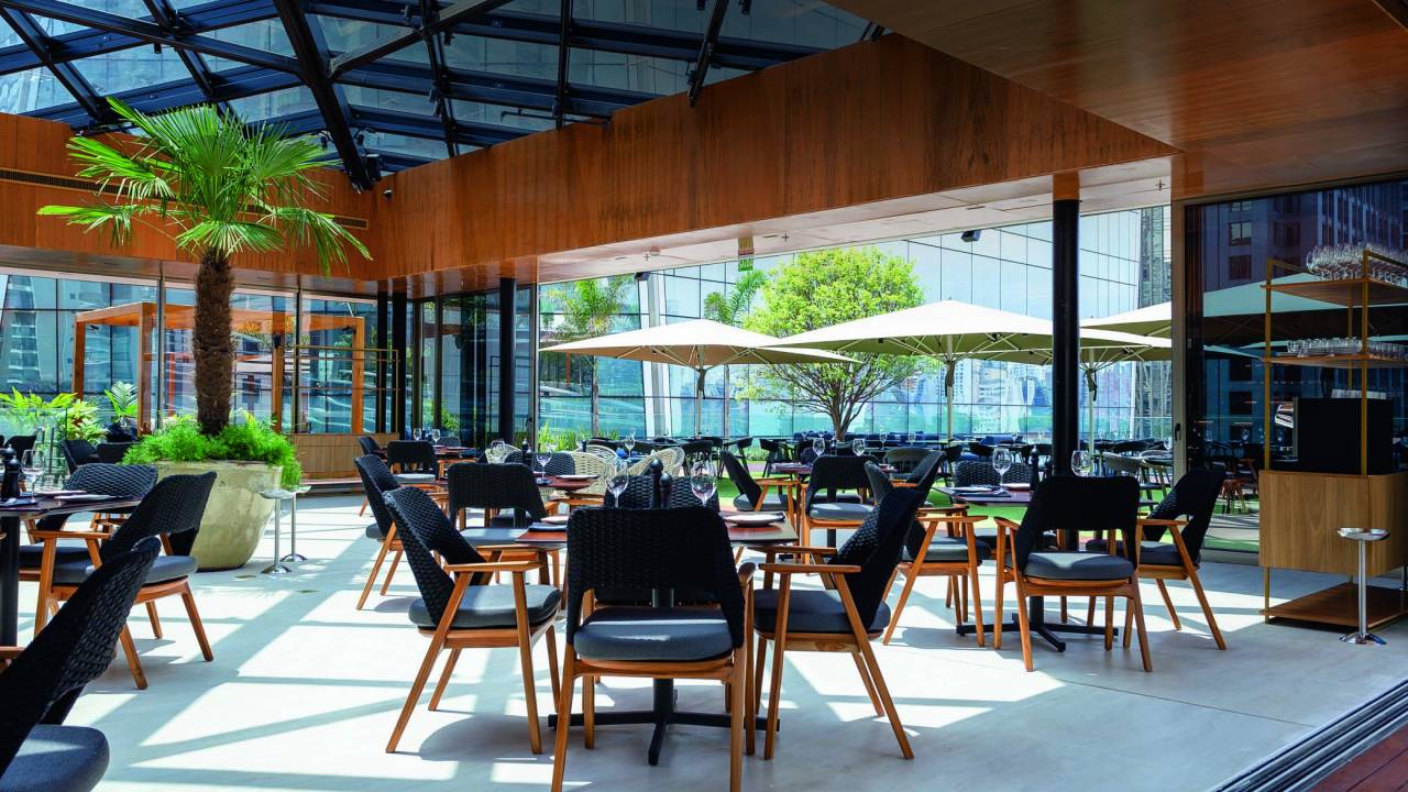 Imagem de ambiente coberto com janelas amplas envidraçadas e mesas com cadeiras e ombrelones