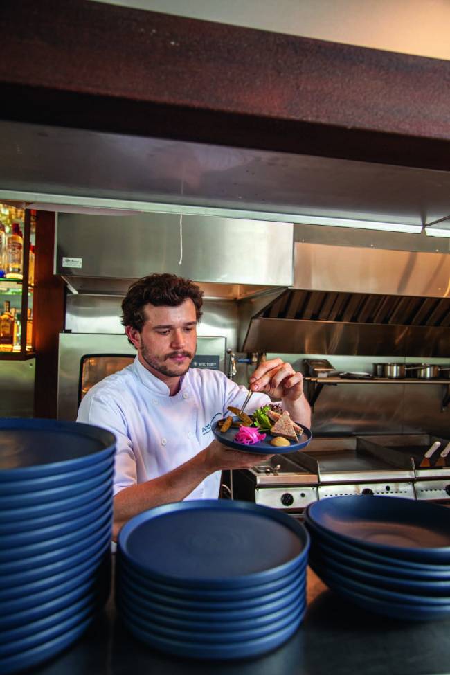 Lucas Bassoleil Avec Gastronomie Homem em uma cozinha com revestimento de inox de jaleco branco finaliza um prato em um recipiente de louça azul