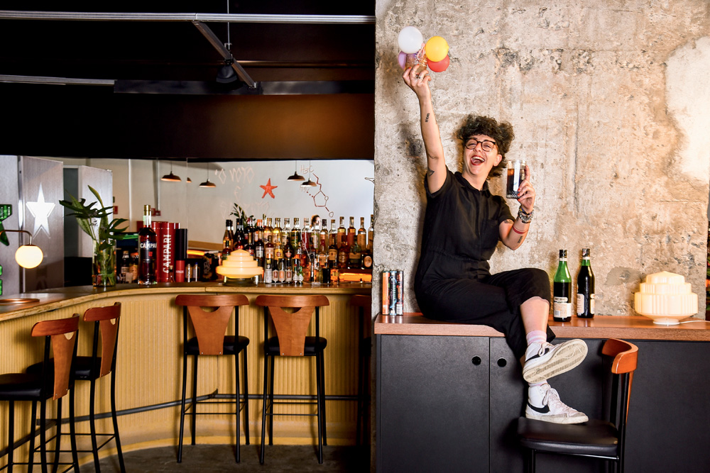 A bartender Chula, uma mulher branca, magra e de cabelos castanhos e cacheados em um corte mullet. Está sentada em uma aparador de bar e segura dois drinques, um em cada mão. Eleva um dos braços e sorri. Usa roupa preta e tênis. Ao fundo, é possível ver um galpão de bar com cadeiras