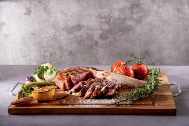 Prime rib com osso maturado – Cór Tábua de madeira com corte de carne maturada acompanhado por vegetais grelhados