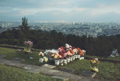 Imagem mostra cemitério com flores em túmulo, em primeiro plano, e uma cidade ao fundo