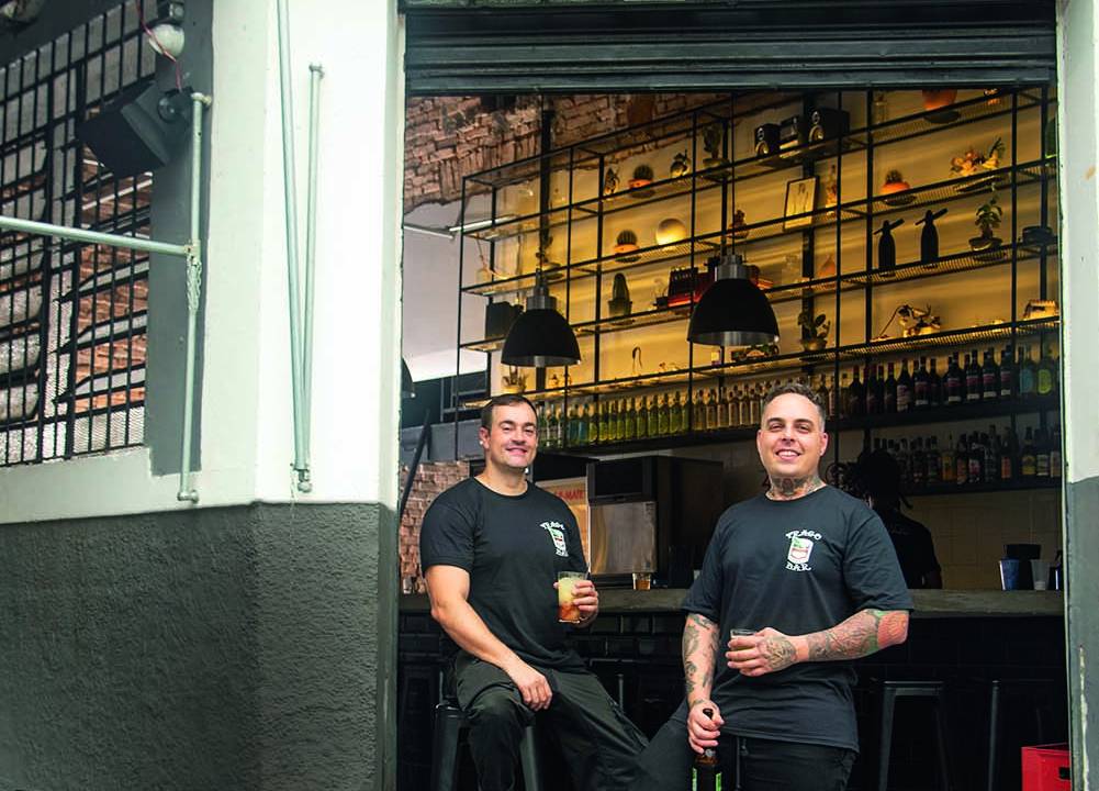 Dois homens vestindo preto em frente a uma fachada aberta sobre a qual se lê o nome estrago em uma placa de madeira amarela