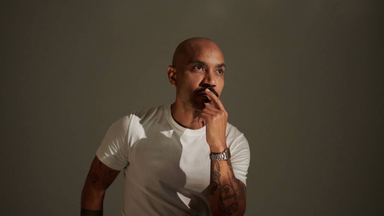 Estilista Luiz Cláudio sentado com uma mão no queixo. Careca de pele negra, veste camiseta branca e calça preta.