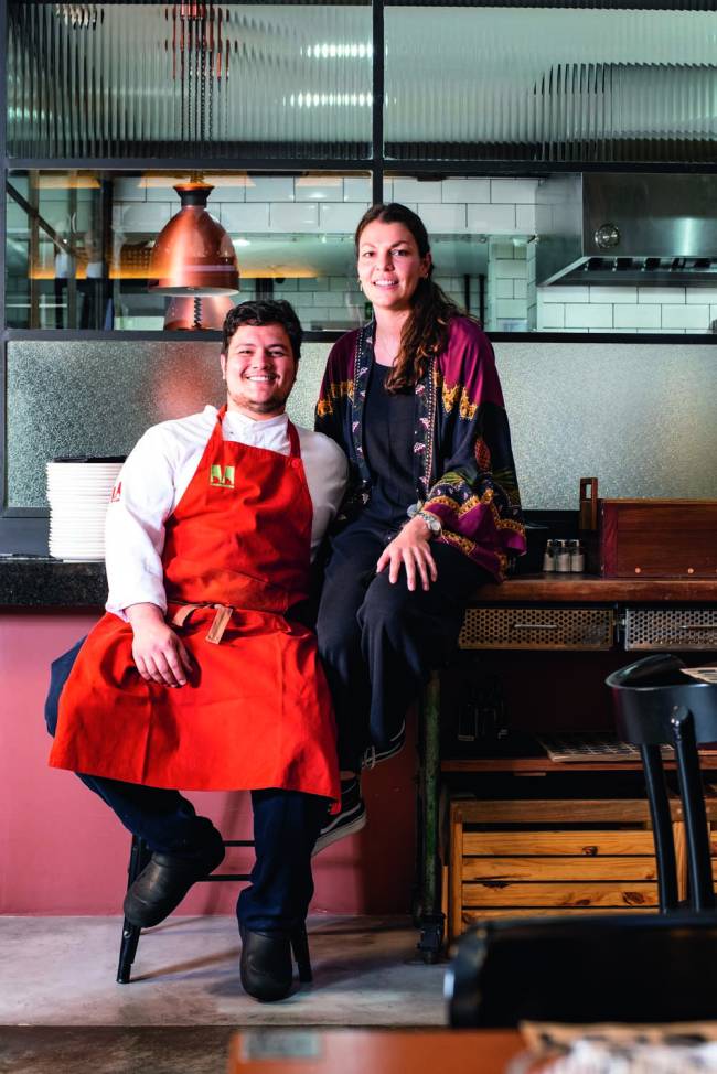 Victor Magri e Charlotte De Cort – Motique Casal com homem vestindo avental vermelho e sentado sobre balcão em ambiente do restaurante