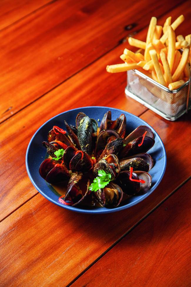 Moules frites Avec Gastronomia Mesa de madeira com tigela azul na qual há mexilhões cozidos na casca e ao lado um recipiente de metal com batatas fritas
