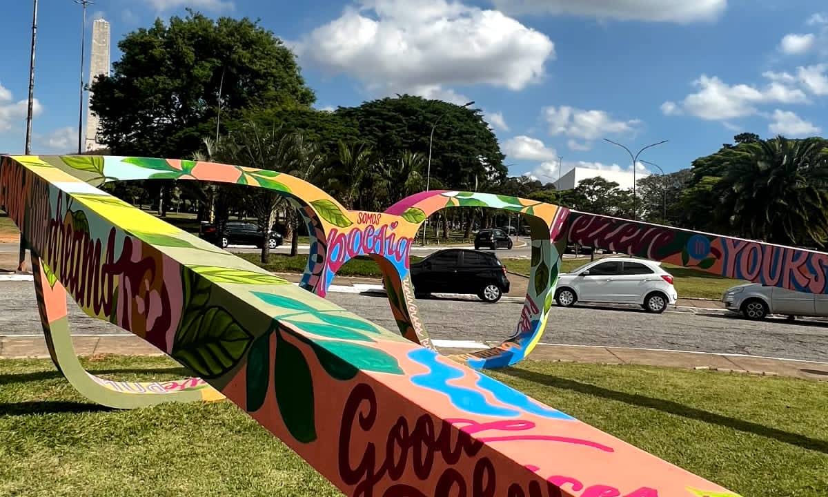 Em gramado, foto exibe par de óculos coloridos gigantes com céu repleto de nuvens.