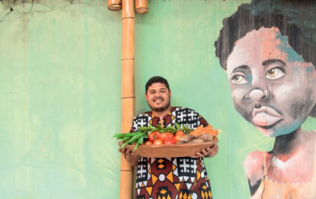 Thiago Vinicius – Organicamente Rango Thiago Vinícius à frente de uma parede na qual há um grafitti de uma mulher negra de blusa laranja, usa uma camiseta de padrão colorido, segura uma cesta de legumes e sorri
