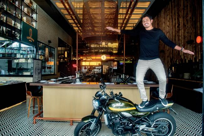 Guilherme Temperani -restaurateur Homem de calça clara e blusa preta de mangas compridas em cima de uma moto preta e amarela com os braços abertos dentro de um espaço interno de restaurante com paredes feitas de bambu e