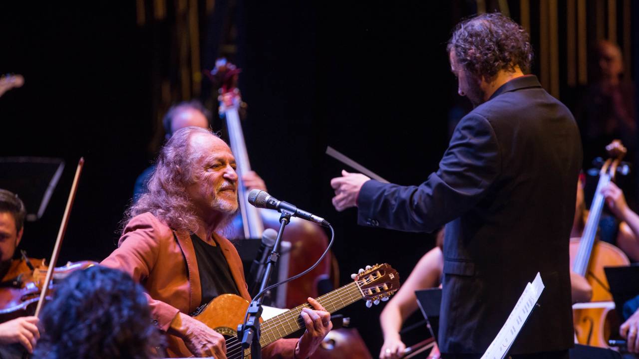 Imagem mostra homem tocando violão, sorrindo, rodeado por músicos de orquestra