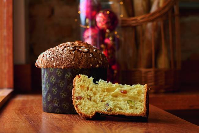 Zestzing Panetone Panetone inteiro ao lado de fatia do panetone cortada, ambos sobre mesa de madeira, com enfeites de natal vermelhos ao fundo