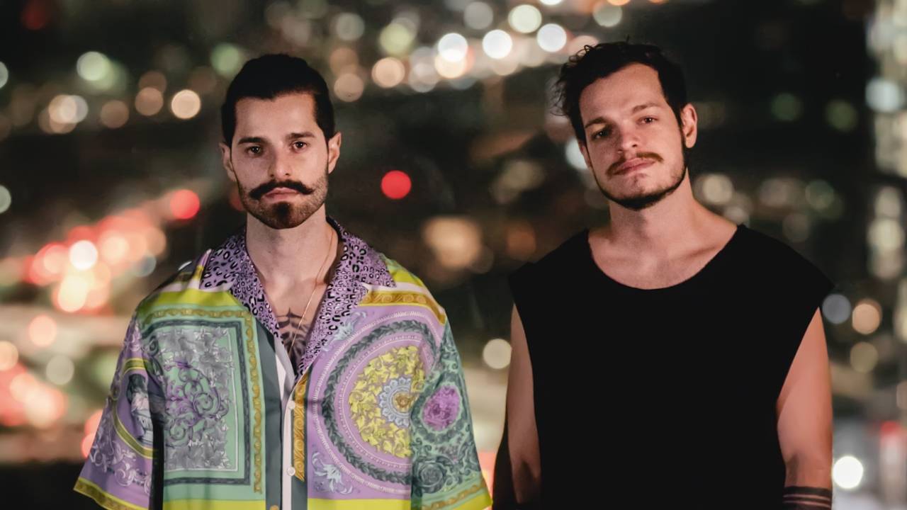 Alok e Bhaskar posam de pé lado a lado. Alok veste camisa estampada e Bhaskar veste regata preta. Ao fundo, luzes da cidade à noite.