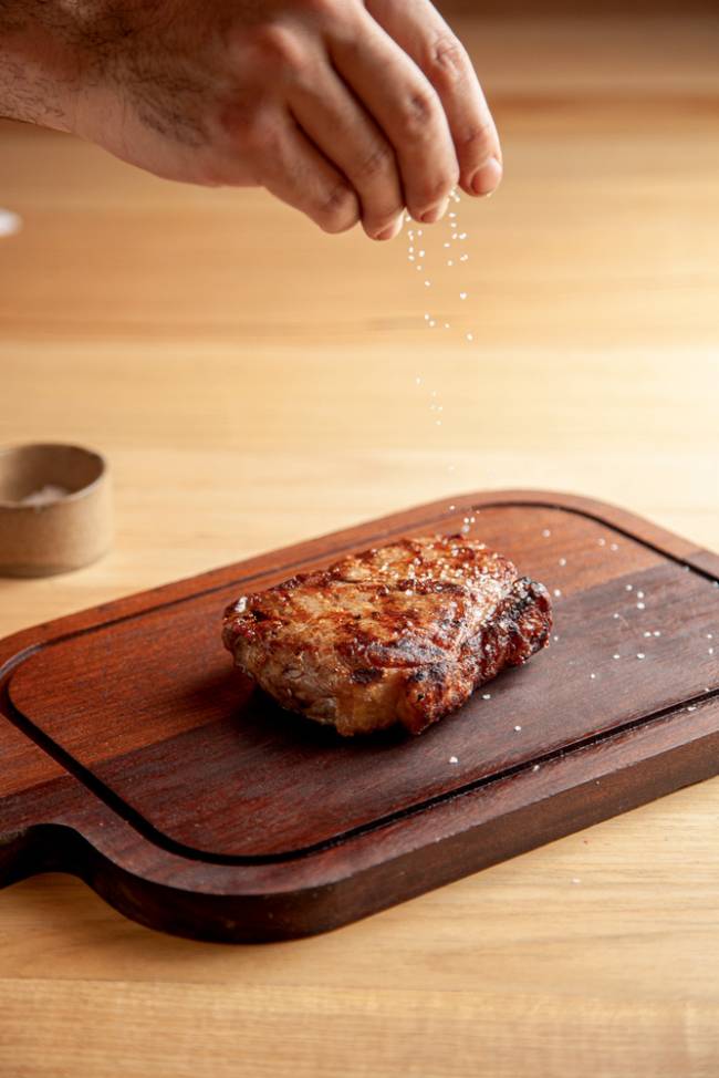 Bife de chorizo do Osso bife de chorizo apoiado sobre tábua de carne em mesa de madeira clara, com mão salpicando sal sobre a peça de carne assada